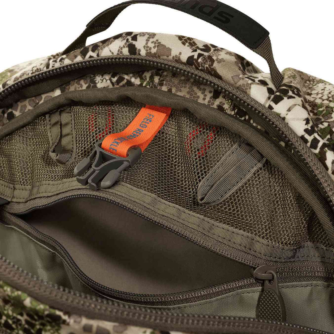 Close-up of a Badlands camo backpack pocket with mesh organizer, zippers, and signature skull logo on the orange key clip.