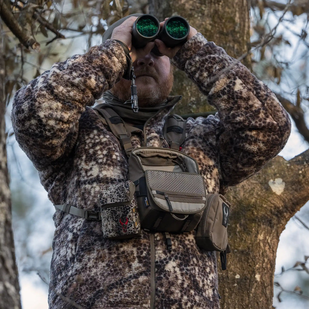 Hunter in camo fleece uses binoculars; Badlands logo skull visible on chest pouch in wooded setting.