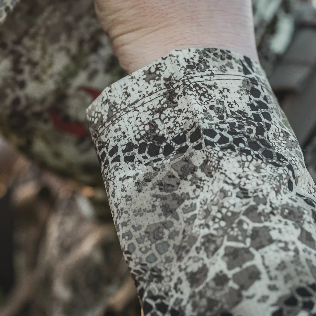 Close-up of a wrist in Badlands camo clothing; blurred Badlands skull logo visible in the background.