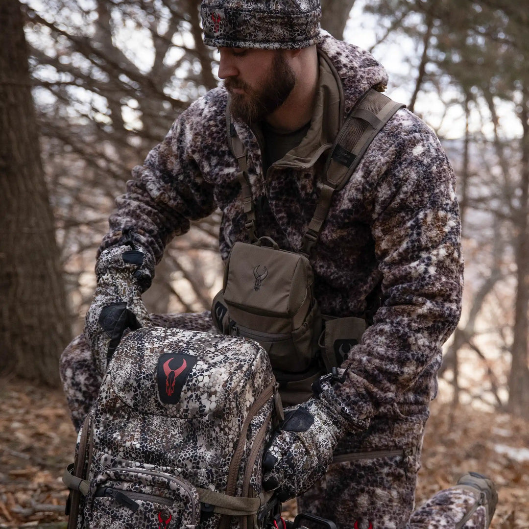 Hunter in Badlands camo gear kneels in the woods with a camo backpack featuring the Badlands skull logo and chest pack.