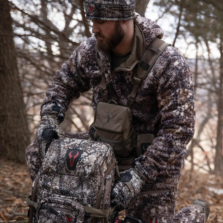 Hunter in Badlands camo gear kneels in the woods with a camo backpack featuring the Badlands skull logo and chest pack.