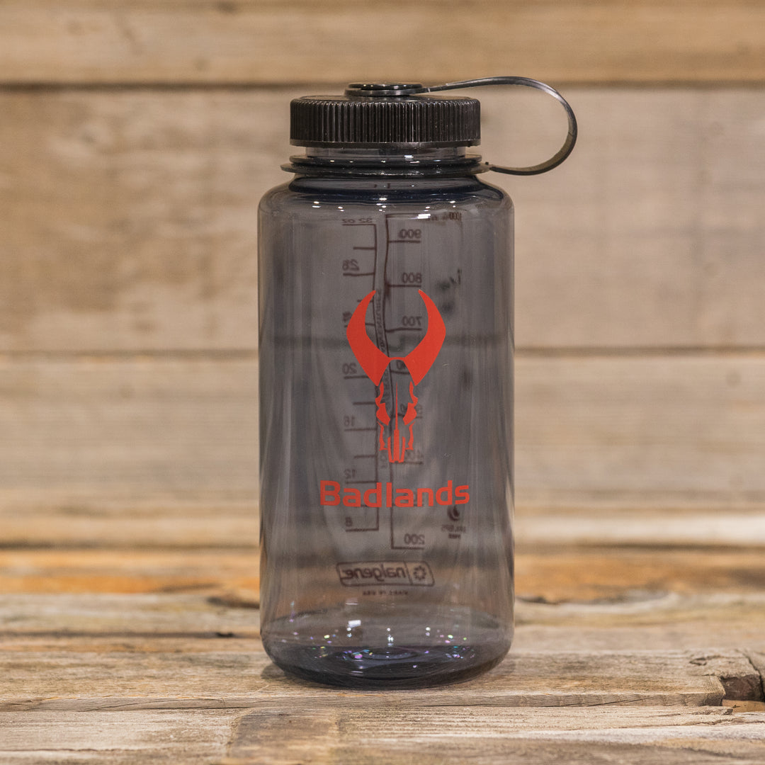 Clear water bottle with black lid, red Badlands logo (skull), and measurement markings; placed on a wooden surface.