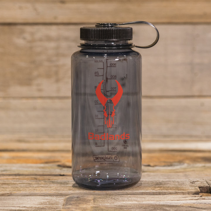 Clear water bottle with black lid, red Badlands logo (skull), and measurement markings; placed on a wooden surface.